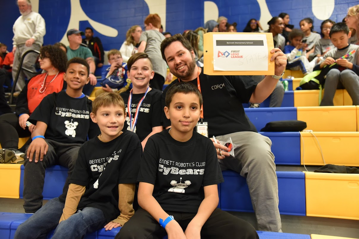students sitting on bleachers with robotics coach holding award certificate