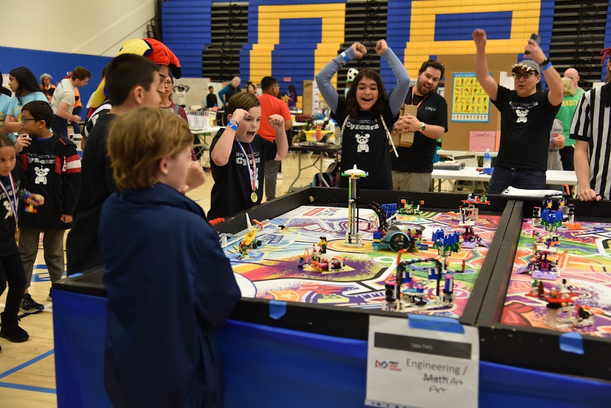 elementary school students standing around a table and cheering at a robotics competition